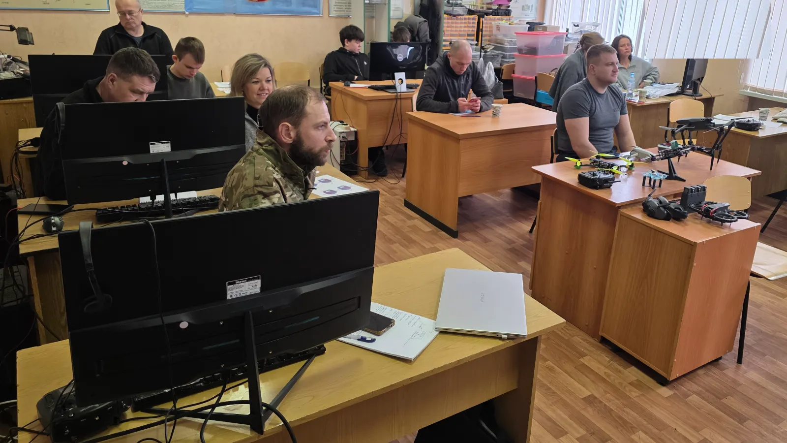 Участники в учебном классе за симуляторами, на столе учебные дроны, пульты и FPV-очки